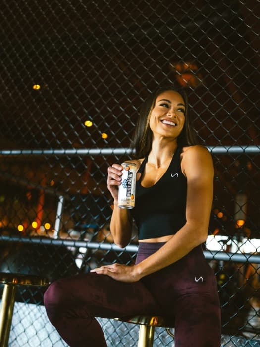 A woman seated on a stool beside a chain-link fence at night with a can of White Rabbit Energy