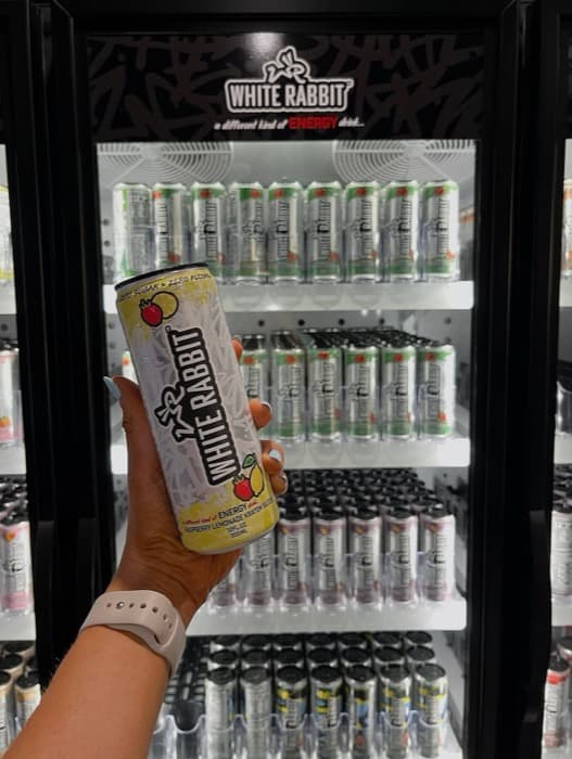 A hand holding a can of White Rabbit Energy in front of a stocked retail cooler